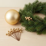 Gold berry picks rest on pale wood beside a gold bauble and a green wreath decorated with another matching pick.