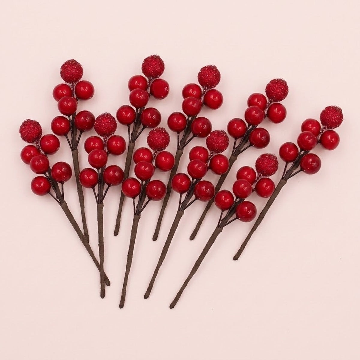 Red foam berry picks with glossy and glittered berries on brown stems fan across a pale pink background.