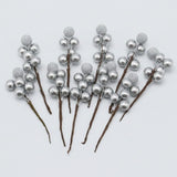 Silver foam berry picks with smooth and glittered berries on brown stems fan across a light grey background.