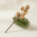 A gold berry pick with a brown wrapped stem rests diagonally across an evergreen sprig on cream fabric.