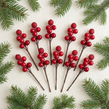 Ten red berry picks with brown stems fan across cream fabric, framed by green evergreen sprigs.