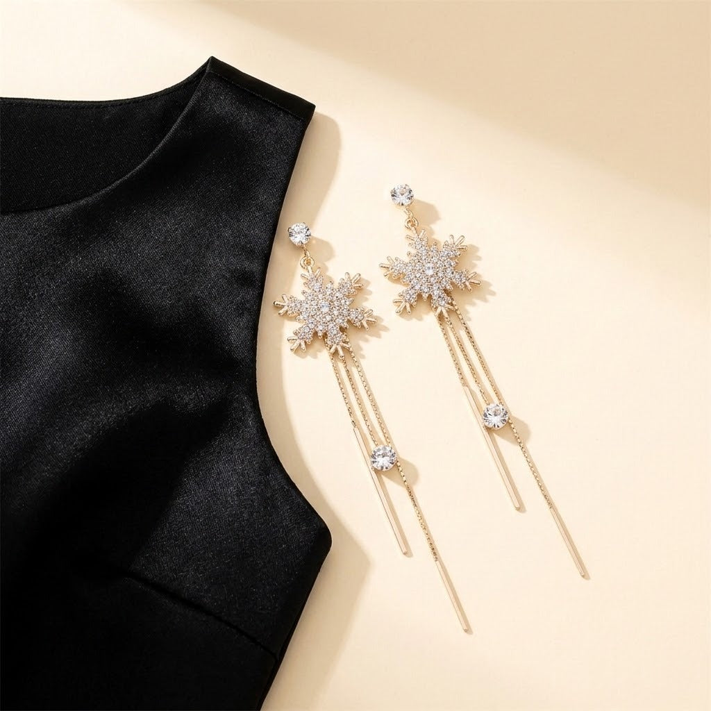 Gold-toned snowflake drop earrings with clear rhinestones and long chain tassels lie beside black fabric on a cream background.