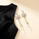 Gold-toned snowflake drop earrings with clear rhinestones and long chain tassels lie beside black fabric on a cream background.