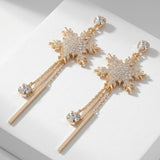 Gold-toned snowflake drop earrings with clear rhinestones, dangling chains and slim bars rest diagonally on a white display block.