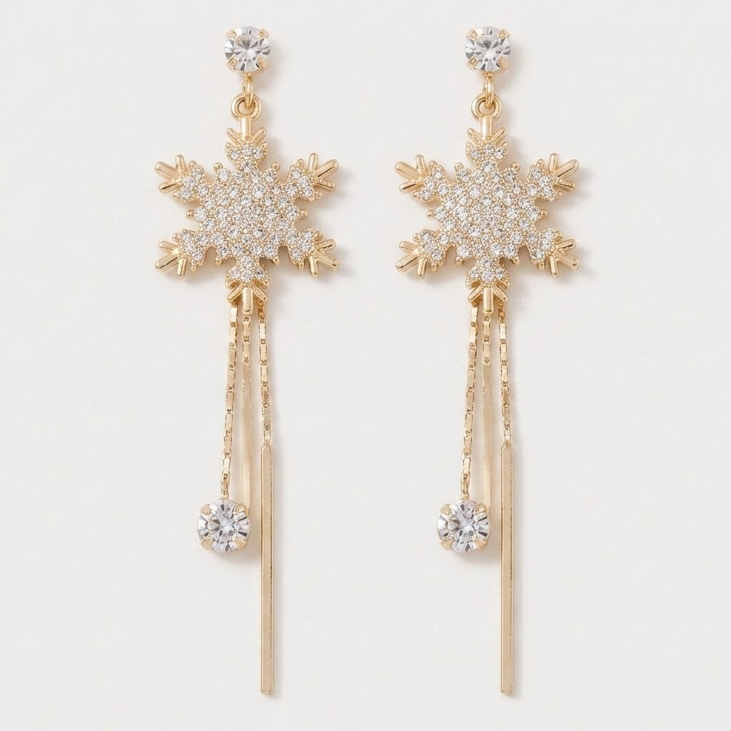 A pair of gold-toned snowflake dangle earrings adorned with clear crystals, featuring a detailed snowflake motif, hanging chains, and round crystal drops on a light off-white background.