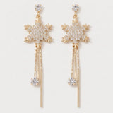 A pair of gold-toned snowflake dangle earrings adorned with clear crystals, featuring a detailed snowflake motif, hanging chains, and round crystal drops on a light off-white background.