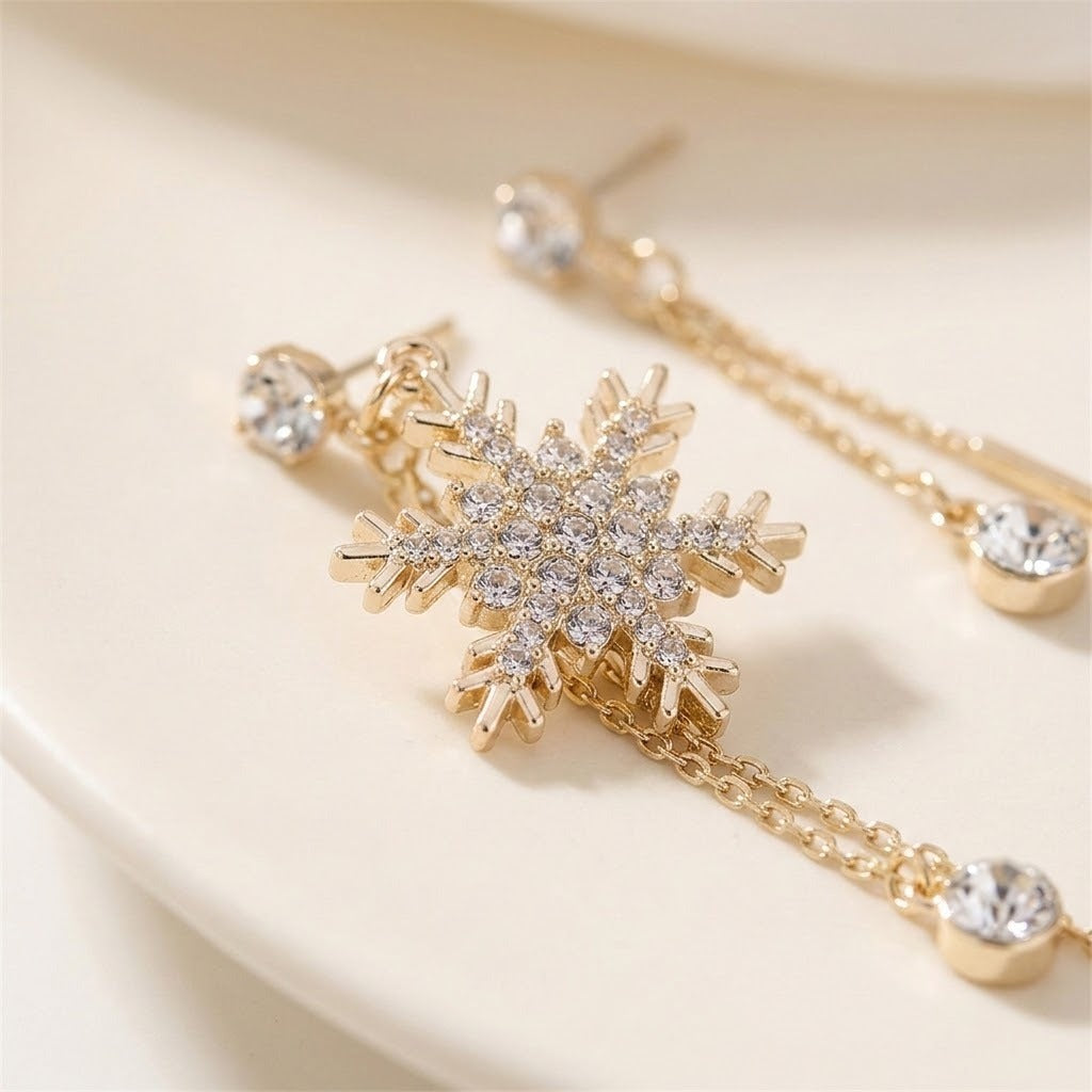 Gold snowflake stud earrings with clear crystal rhinestones and matching drop chains, displayed on a light cream surface.