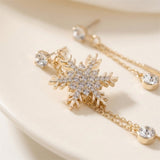 Gold snowflake stud earrings with clear crystal rhinestones and matching drop chains, displayed on a light cream surface.