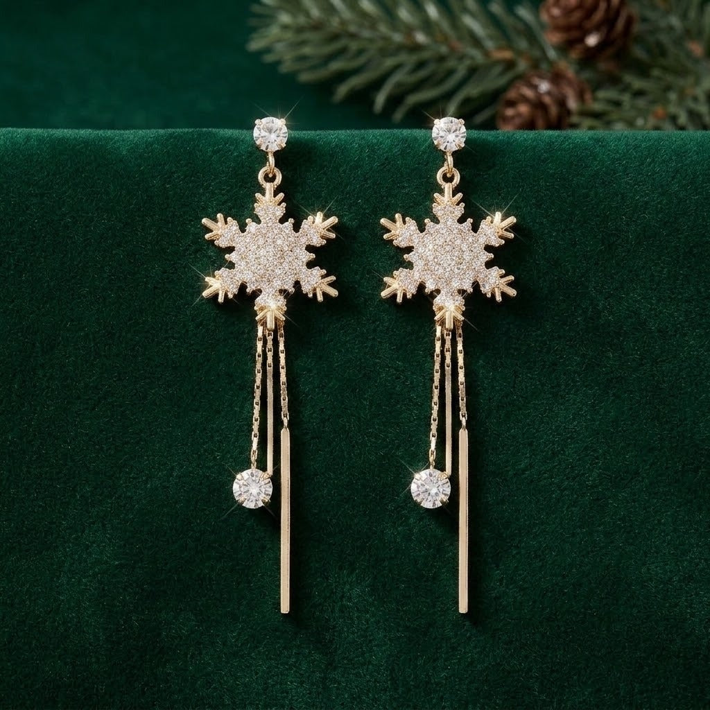 A pair of gold-toned snowflake dangle earrings with clear crystal embellishments and long vertical chain drops, displayed on a dark green velvet surface with pine branches in the background.
