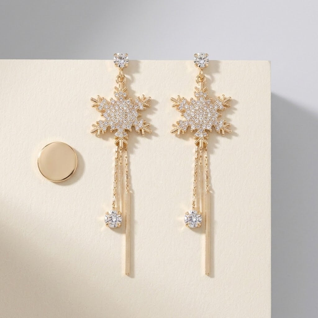 Gold-toned snowflake drop earrings with clear rhinestones and chain tassels rest on a cream card beside a round gold-toned disc.