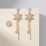 Gold-toned snowflake drop earrings with clear rhinestones and chain tassels rest on a cream card beside a round gold-toned disc.