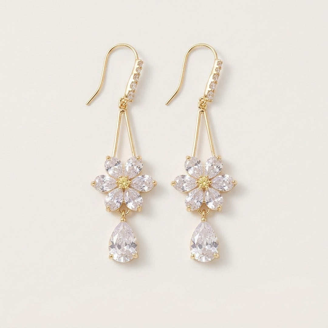 Gold-toned drop earrings with clear rhinestone flowers, teardrop pendants and curved ear hooks lie against an off-white background.