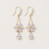 Gold-toned drop earrings with clear rhinestone flowers, teardrop pendants and curved ear hooks lie against an off-white background.