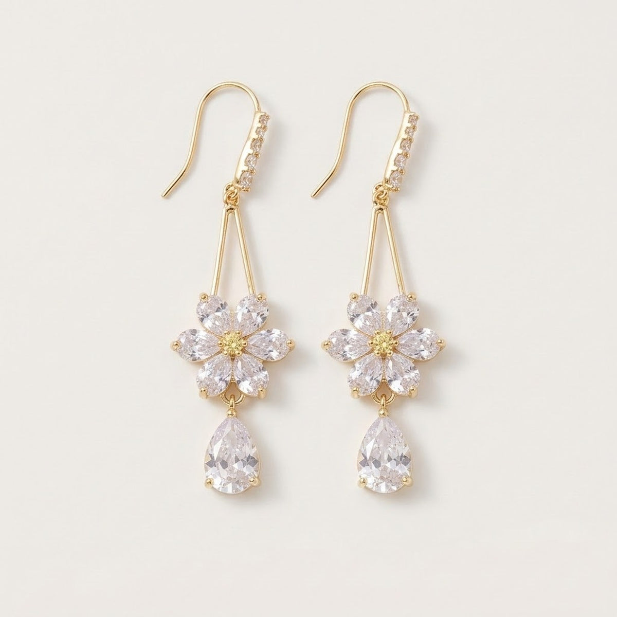 Gold-toned drop earrings with clear rhinestone flowers, teardrop pendants and curved ear hooks lie against an off-white background.