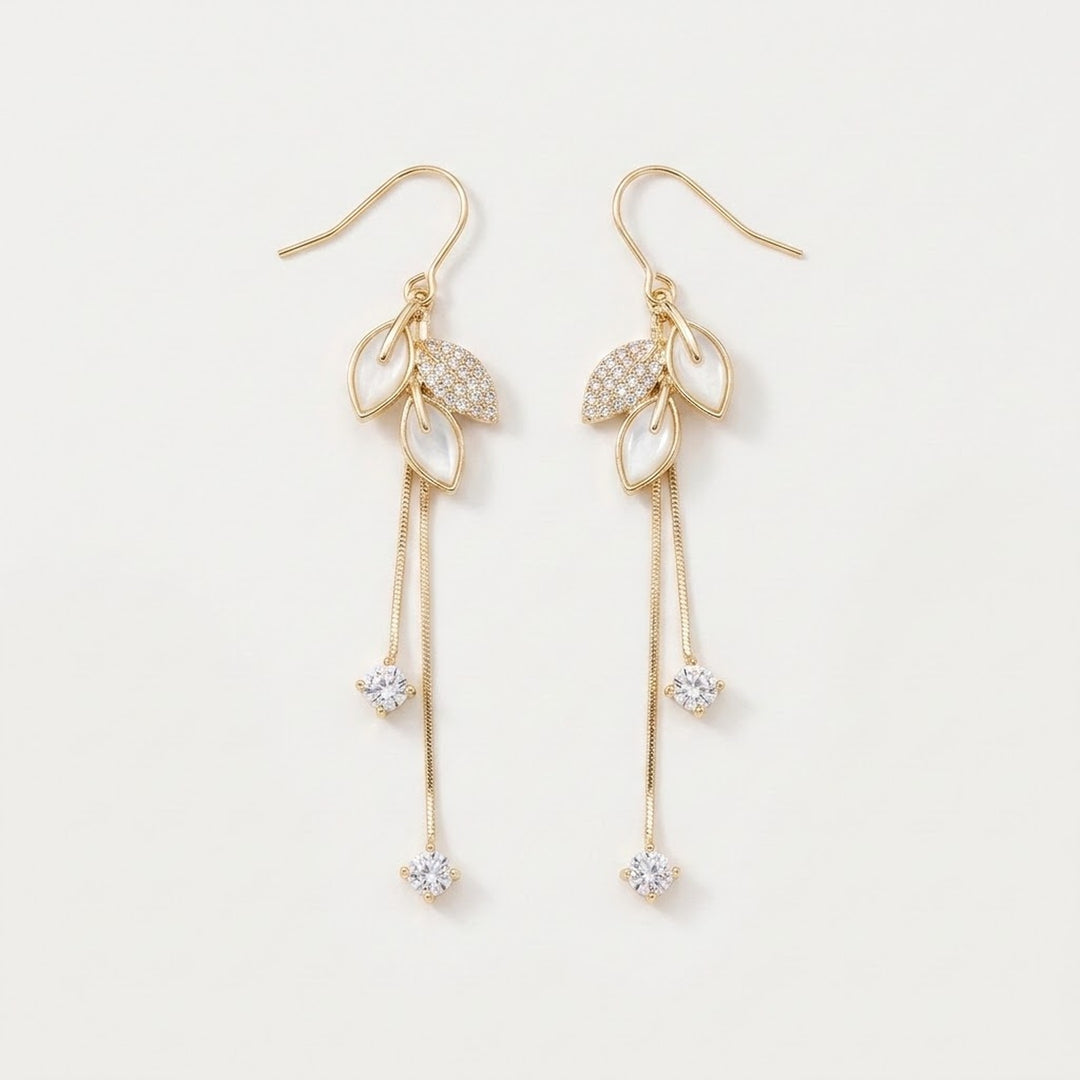Gold-tone drop earrings with white leaf-shaped accents, clear rhinestones, and long chain tassels lie on a white background.