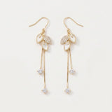 Gold-tone drop earrings with white leaf-shaped accents, clear rhinestones, and long chain tassels lie on a white background.
