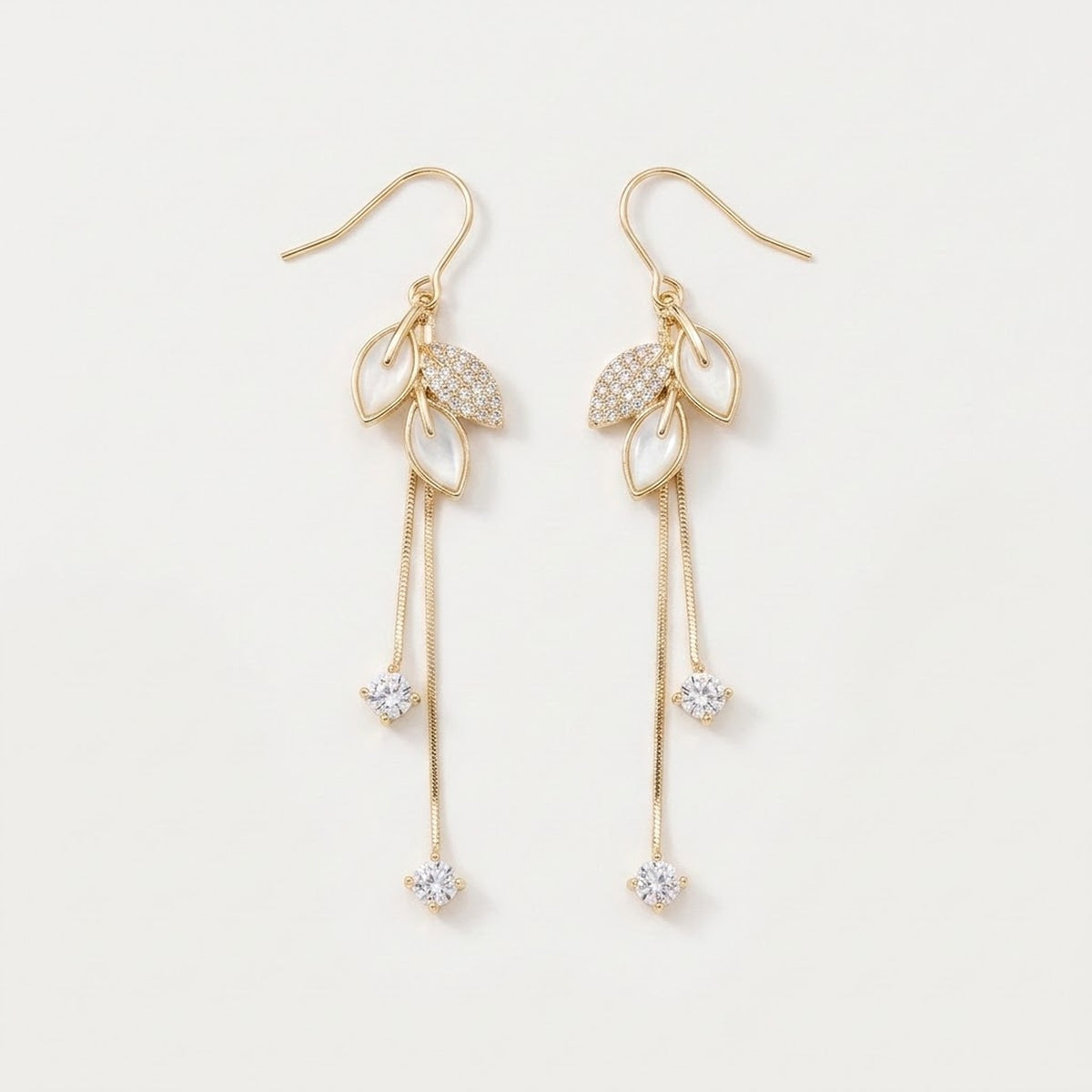 Gold-tone drop earrings with white leaf-shaped accents, clear rhinestones, and long chain tassels lie on a white background.
