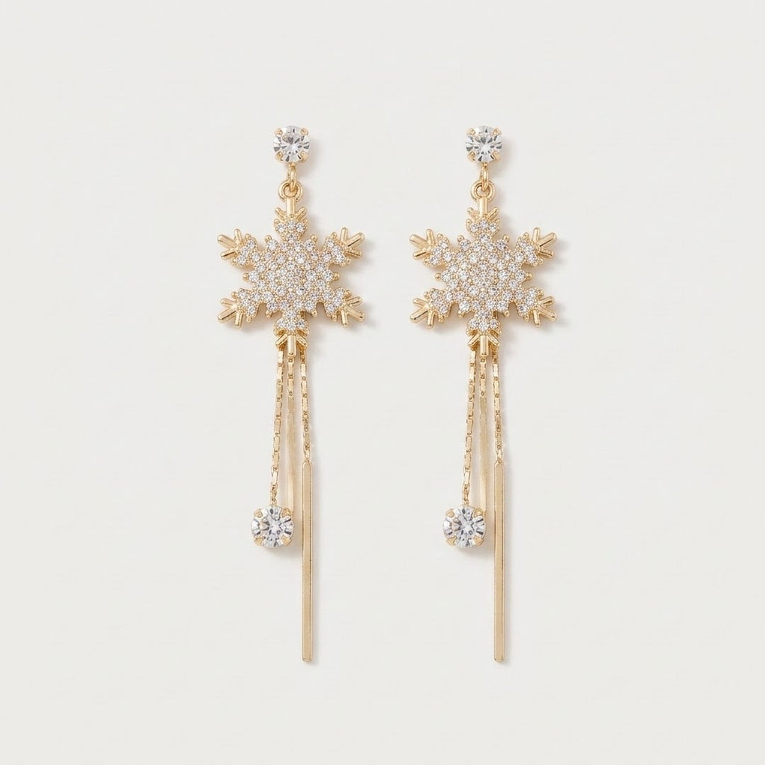 Gold-tone drop earrings with rhinestone snowflakes, dangling chains, clear stones, and slender bar tassels lie on a white background.