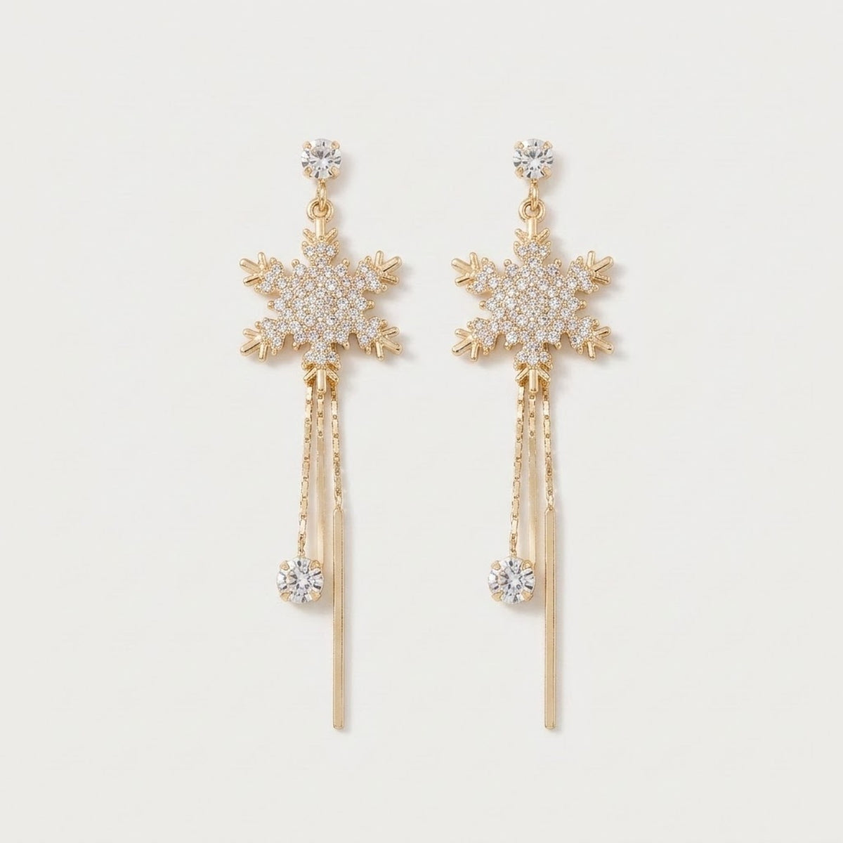 Gold-tone drop earrings with rhinestone snowflakes, dangling chains, clear stones, and slender bar tassels lie on a white background.