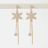 Gold-toned snowflake drop earrings with clear rhinestones and long dangling bars hang from a white display stand.