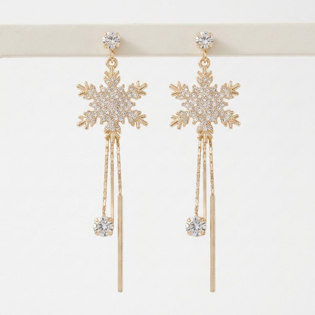 Gold-toned snowflake drop earrings with clear rhinestones and long dangling bars hang from a white display stand.