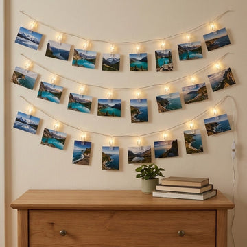 Warm white photo clip lighting holds landscape prints in three curved rows on a beige wall above a wooden dresser.