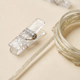 Photo clip lighting close-up shows a clear plastic clip with a metal spring beside coiled transparent cord on a beige background.