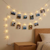 Warm white photo clip lighting displays landscape and pet photographs on a beige bedroom wall above a wooden nightstand beside the bed.