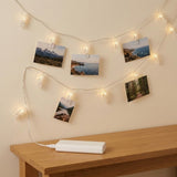 Warm white photo clip lighting holds landscape photographs on a cream wall above a wooden table with a white power bank.