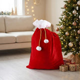 Red gift sack with white trim, gold drawstrings, and white pom-poms stands beside a decorated Christmas tree and wrapped presents.
