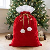 Red gift sack with white trim and gold cords tied in a bow stands before a Christmas tree with presents and pinecones.