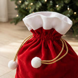 Close-up of a red gift sack shows gathered fabric, white trim, gold drawstrings, and white pom-poms against a blurred Christmas tree.