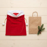 Red fabric gift sack with white trim and pompoms beside a brown paper bag and evergreen sprig on pale wooden boards.
