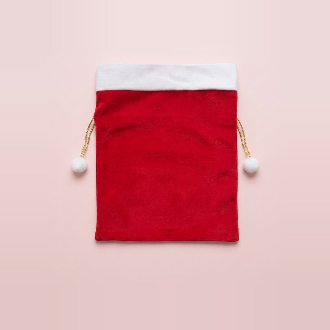 Red fabric gift sack with white trim and gold drawstrings tipped with white pompoms sits centered against a pale pink background.