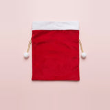 Red fabric gift sack with white trim and gold drawstrings tipped with white pompoms sits centered against a pale pink background.