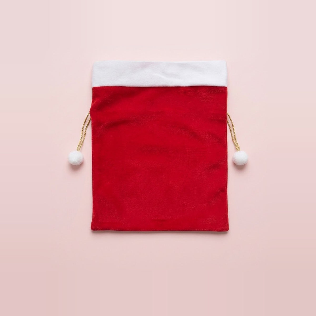 Red fabric gift sack with white trim and gold drawstrings tipped with white pompoms sits centered against a pale pink background.