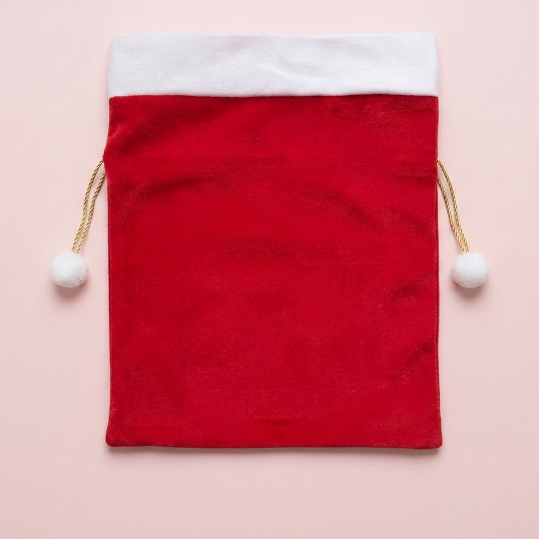Red fabric gift sack with a white cuff and gold drawstrings ending in white pompoms lies flat on a pale pink background.