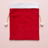 Red fabric gift sack with a white cuff and gold drawstrings ending in white pompoms lies flat on a pale pink background.