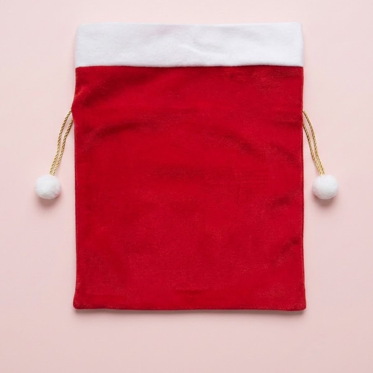 Red fabric gift sack with a white cuff and gold drawstrings ending in white pompoms lies flat on a pale pink background.