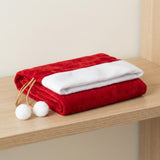 Folded red gift sack with white trim, gold cords, and two white pom-poms rests on a light wooden shelf.