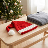 A red knit Santa hat with a white cuff and white pom-pom rests on a light wood table next to a folded gray sweater, with a softly blurred Christmas tree in the background.