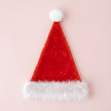 A red Santa hat with a fuzzy white cuff and round white pompom lies flat on a pale pink background.