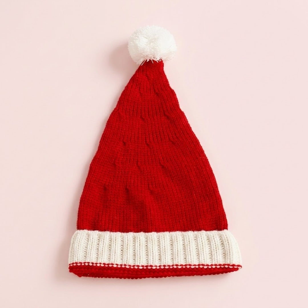 A red knitted Santa hat with a white ribbed cuff and fluffy white pompom lies flat on a pale pink background.