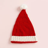 A red knitted Santa hat with a white ribbed cuff and fluffy white pompom lies flat on a pale pink background.