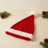 A red knitted Santa hat with a white ribbed cuff and pompom rests on pale wood among evergreen sprigs and pinecones.