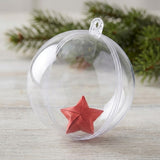 A clear plastic fillable ornament holds a folded red star on pale wood with evergreen foliage behind it.