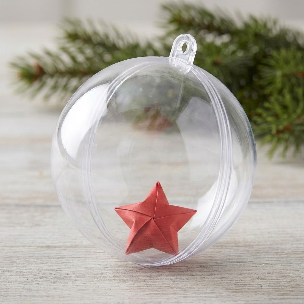 A clear plastic fillable ornament holds a folded red star on pale wood with evergreen foliage behind it.