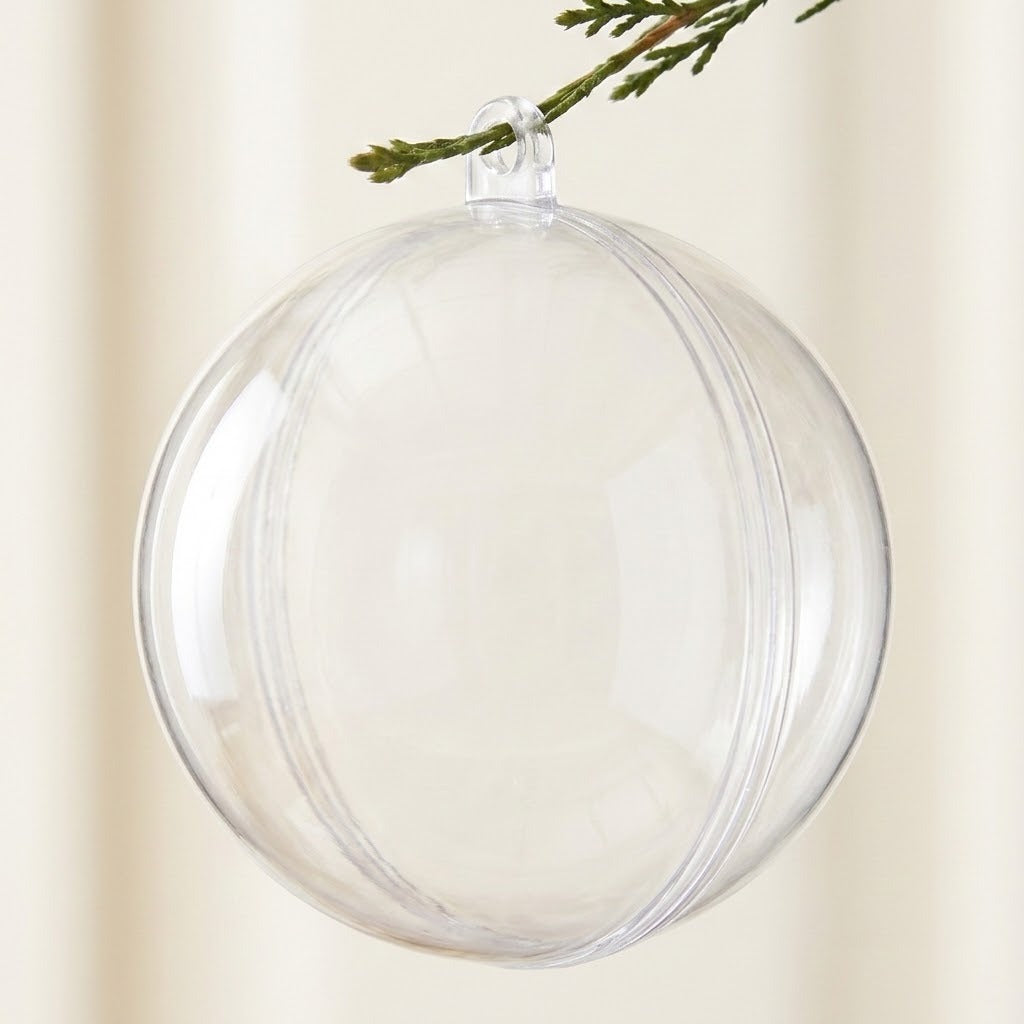 A clear plastic fillable ornament hangs by its top loop from an evergreen twig against a cream background.