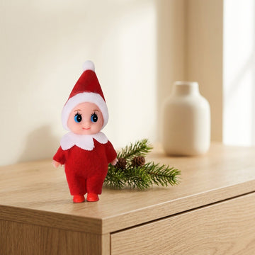 A red Christmas elf doll with white trim stands on a wooden cabinet beside evergreen sprigs, with a cream vase behind.