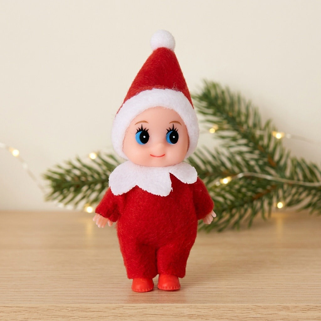 A blue-eyed Christmas elf doll in red fabric clothing stands on a wooden surface before evergreen branches and warm fairy lights.